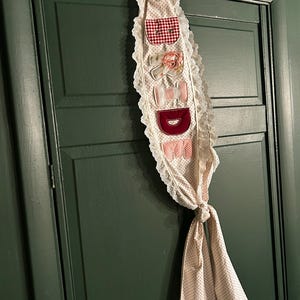 May include: A decorative door hanging with the letters "BED" stitched onto it. The hanging features pockets and bows in red gingham, floral, and pink polka dot patterns, trimmed with lace. The hanging is tied at the bottom with a matching polka dot fabric.