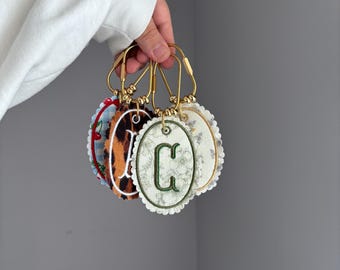 May include: A collection of four oval-shaped fabric keychains with scalloped edges, each featuring a different pattern including cherries, leopard print, and floral designs. Each keychain is attached to a gold-toned metal ring with a letter monogram.