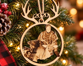 May include: A circular wooden Christmas ornament featuring a laser-engraved image of a person with a deer and a bow. The top of the ornament is shaped like deer antlers with the year 2020 cut out. It hangs from a jute twine loop.