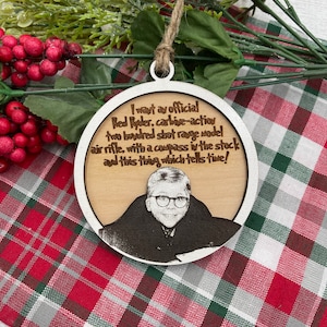 May include: A round wooden Christmas bauble with a laser-engraved image of a man and text that reads: "I want ay official Red Ryder, carbive-action two buvored shot range model air rifle, with a compass in the stock and this thing which tells time!"