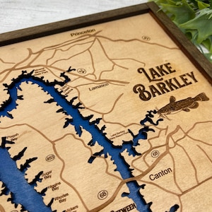 Op de afbeelding: Een houten kaart van Lake Barkley, Kentucky, met de omtrek van het meer in blauw en het omliggende land in bruin. De kaart bevat de namen van verschillende baaien en inhammen, evenals de tekst "Land Between The Lakes".