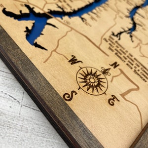 Op de afbeelding: Een houten kaart met een uitgegraven blauw meer, omgeven door een donkerbruine lijst. De kaart heeft een kompas met de richtingen Noord, Zuid, Oost en West. De tekst op de kaart luidt "This beauty is the westernmost point of the Shenandoah River. You can't go alone in a canoe. One must paddle with a companion. You paddle with Kentucky."