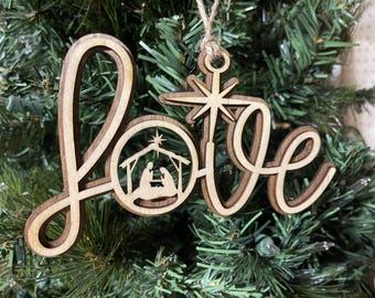 Puede incluir: Adorno navideño de madera con la palabra "love" en una fuente cursiva. El adorno presenta un recorte de la escena de la natividad con una estrella sobre el establo.
