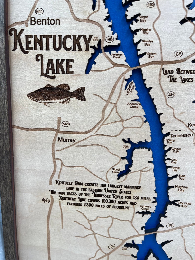Op de afbeelding: Een houten kaart van Kentucky Lake met een blauwe omtrek van het meer en het omliggende land. De kaart bevat de namen van verschillende baaien en kreken, evenals de tekst "Kentucky Dam cre&euml;ert het grootste kunstmatige meer in het oosten van de Verenigde Staten. De dam stuwt de Tennessee River 184 mijl op. Kentucky Lake beslaat 160.300 acres en heeft 2.300 mijl kustlijn."