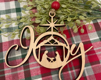 Puede incluir: Adorno navideño de madera con la palabra "Joy" en escritura cursiva. El adorno presenta una silueta de la escena de la natividad dentro de un círculo.