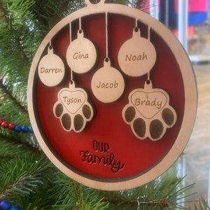 May include: A round wooden Christmas ornament with a red background. The ornament features the names "Darren", "Gina", "Noah", "Jacob", "Tyson", and "Brady" in a decorative font. The ornament also includes the words "Our Family" in a cursive font.