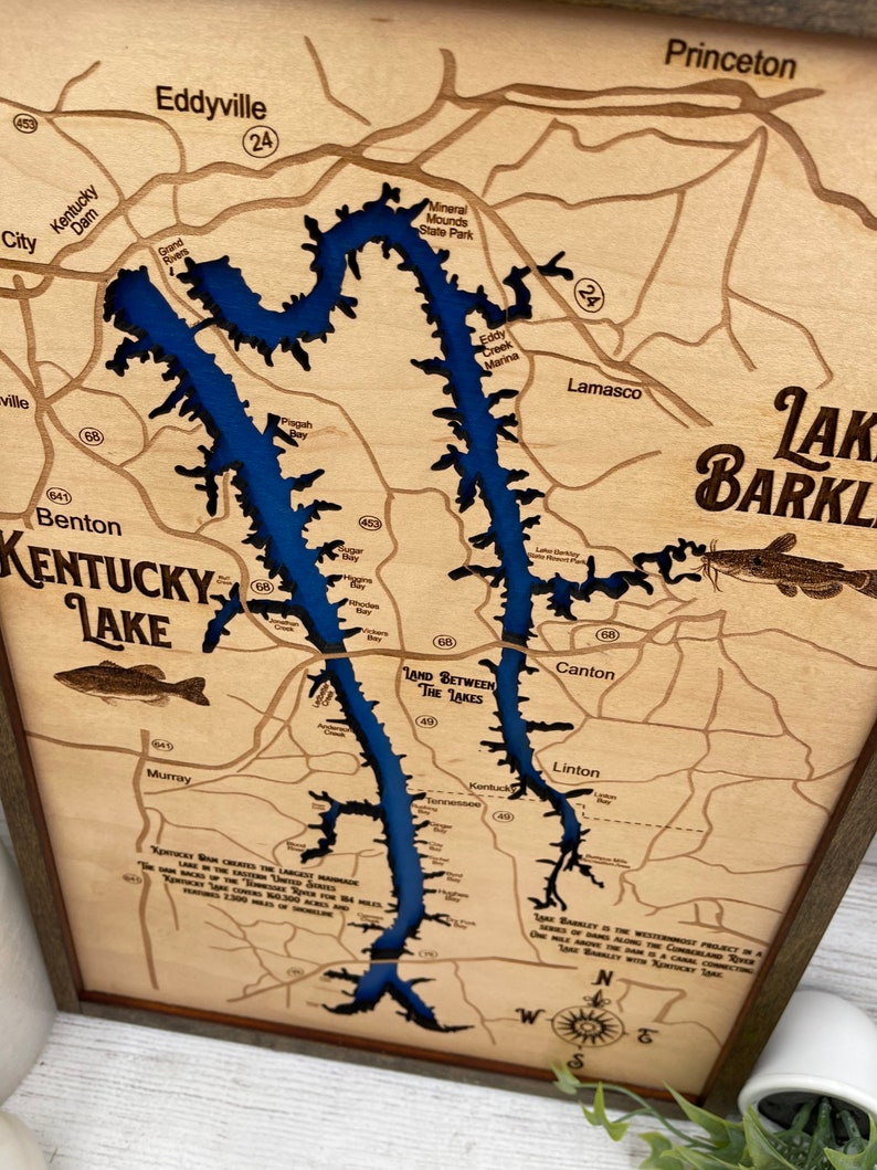 Op de afbeelding: Een houten kaart van Lake Barkley in Kentucky, met de omtrek van het meer uitgesneden en blauw geverfd. De kaart bevat de namen van steden en bezienswaardigheden rond het meer, zoals Eddyville, Benton en Princeton. De tekst "Kentucky Lake" staat prominent linksonder op de kaart. De tekst "Land Between The Lakes" staat in het midden van de kaart. De tekst "Lake Barkley is the westernmost project in a series of dams along the Cumberland River. One mile above the dam is a connecting river, Lake Barkley within Kentucky Lake." staat rechtsonder op de kaart.