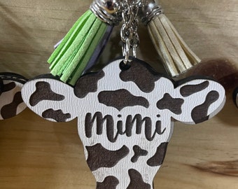 May include: Cow-print keychain with a black and white cow design and the name "mimi" engraved on it. The keychain has two tassels, one green and one white.