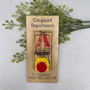 May include: A wooden sign with the text "Complaint Department" and a mouse trap with a red button. The text below the trap reads "To complain... PUSH RED BUTTON".