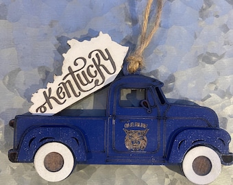 May include: A blue wooden truck ornament with a white outline and a white wooden cutout of the state of Kentucky with the word "Kentucky" written in cursive on it. The truck has a white tire on each side and a Wildcat logo on the side that says "Wildcats".