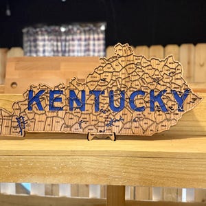 May include: Wooden Kentucky state map with blue lettering spelling "Kentucky". The map is divided into counties, with each county labeled. The map is displayed on a wooden shelf, with a black background.