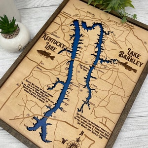 Op de afbeelding: Een houten kaart van Lake Barkley in Kentucky, met een blauw meer en bruin land. De kaart is ingelijst in een donkerbruin frame. De tekst op de kaart luidt "Kentucky Dam cre&euml;ert het grootste kunstmatige meer in het oosten van de Verenigde Staten. De dam stuwt de Tennessee River 18 mijl op. Kentucky Lake beslaat 60.300 acres en heeft 2.300 mijl kustlijn." en "Lake Barkley is het meest westelijke project in een reeks dammen langs de Cumberland River. Een mijl boven de dam is er een kanaal dat Lake Barkley verbindt met Kentucky Lake."
