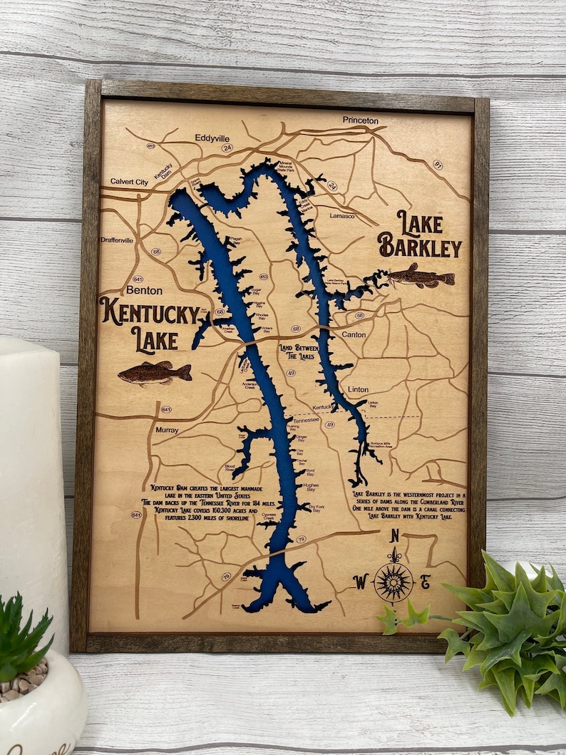 Op de afbeelding: Een houten kaart van Lake Barkley en Kentucky Lake, met een blauwe wateruitsnijding en een bruin houten frame. De kaart bevat de tekst "Kentucky Lake" en "Lake Barkley" samen met de namen van nabijgelegen steden en dorpen. De kaart bevat ook een kompasroos en een beschrijving van de meren.