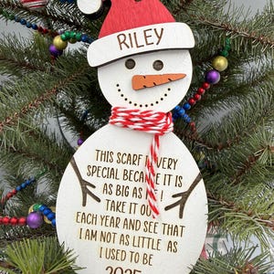 May include: A wooden snowman Christmas ornament with a red hat and scarf. The ornament has the name "RILEY" on the hat and the text "THIS SCARF IS VERY SPECIAL...2025" on the body. It is hanging on a Christmas tree.