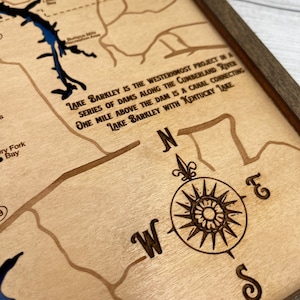 Op de afbeelding: Een houten kaart van een meer met een kompasroos en de tekst "LAKE BARKLEY IS THE WESTERNMOST PROJECT IN A SERIES OF DAMS ALONG THE CUMBERLAND RIVER ONE MILE ABOVE THE DAM IS A CANAL CONNECTING LAKE BARKLEY WITH KENTUCKY LAKE".