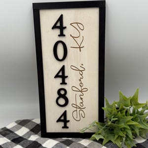 Może przedstawiać: Prostokątny drewniany szyld z adresem z czarną ramą. Szyld wyświetla czarne cyfry "40484", z "KS" i "Stanford" w brązowej czcionce. Tło jest w jasnym kolorze drewna.