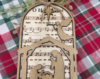 Puede incluir: Un adorno navideño de madera con forma de portal de Belén con un recorte de una estrella y partitura musical. El adorno tiene las palabras "O Holy Night!" y las letras "The stars are brightly shining" y "Long lay the world."