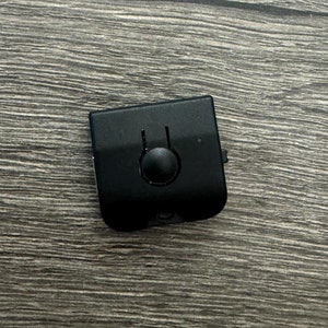 May include: A small, black, square-shaped electronic component with a circular button and two vertical lines. The component is set against a gray wooden surface.