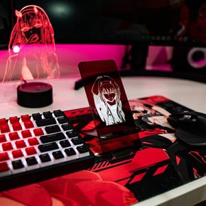 May include: A red and black gaming setup featuring a keyboard with red keycaps, a mouse, and a mousepad with anime graphics. A red acrylic LED lamp and a phone stand with anime art are also present.