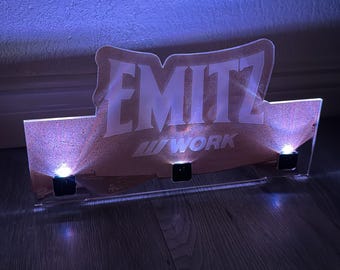 May include: A rectangular acrylic display sign featuring the text EMITZ and WORK in white lettering. The sign is illuminated from below by three small LED lights, casting a blue glow across the surface and the surrounding area.