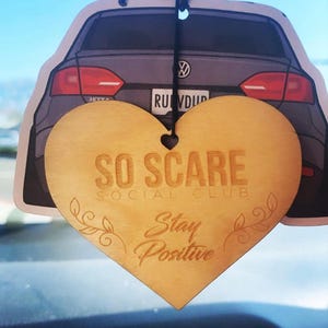 May include: A wooden heart-shaped car air freshener with the text "So Scare Social Club" and "Stay Positive" printed on it. The air freshener is hanging from a rearview mirror in a car.