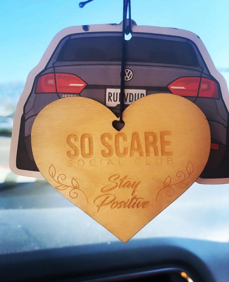 May include: A wooden heart-shaped car air freshener with the text "So Scare Social Club" and "Stay Positive" printed on it. The air freshener is hanging from a rearview mirror in a car.