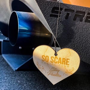 May include: A wooden heart-shaped ornament with the text "SO SCARE SOCIAL CLUB Stay Positive" in gold lettering. The ornament is hanging from a black string.