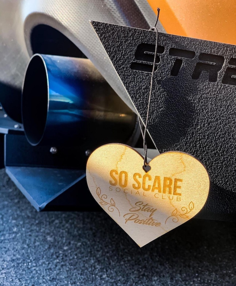 May include: A wooden heart-shaped ornament with the text "SO SCARE SOCIAL CLUB Stay Positive" in gold lettering. The ornament is hanging from a black string.