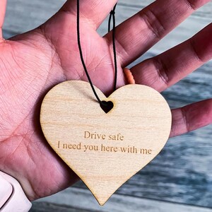 May include: A wooden heart-shaped car air freshener with a black string for hanging. The heart is engraved with the text "Drive safe I need you here with me".