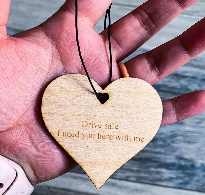 May include: A wooden heart-shaped car air freshener with a black string for hanging. The heart is engraved with the text "Drive safe I need you here with me".