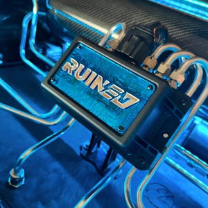 May include: A close-up of a black and blue engine part with the word "RUINED" printed on it. The part is connected to a series of silver pipes and is surrounded by blue lights.