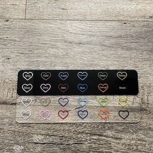 May include: A clear acrylic sheet with a black background and rows of heart-shaped stickers. The stickers are outlined in different colors, including white, gold, purple, turquoise, green, yellow, silver, pink, red, blue, orange, and black.