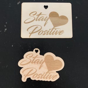 May include: Two wooden ornaments with the text "Stay Positive" and a heart shape. The ornaments are made of light brown wood and have a natural finish.