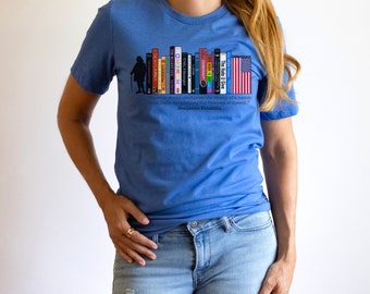 Puede incluir: Camiseta azul con un diseño de estantería, una bandera estadounidense y la silueta de una persona. La cita de Benjamin Franklin, "Quien quiera derrocar la libertad de una nación debe empezar por someter la libertad de expresión", está impresa debajo.