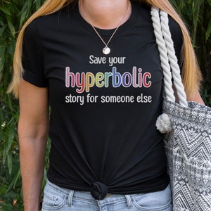 May include: Black t-shirt with the text "Save your hyperbolic story for someone else" in colorful lettering. The shirt is knotted at the waist. A necklace and a patterned tote bag with a white rope handle are also visible.