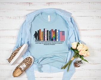 Puede incluir: Camiseta azul claro con un gráfico de libros y una bandera estadounidense, con una cita de Benjamin Franklin. La camiseta está sobre una camisa a rayas azules, con zapatos con estampado de leopardo y flores.