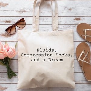 Könnte beinhalten: Eine weiße Canvas-Tragetasche mit dem Text "Fluids, Compression Socks, and a Dream" in schwarzer Schrift. Die Tasche liegt auf einer weißen Holzoberfläche mit rosa Blumen, Sonnenbrillen und zwei Paar goldenen Sandalen.
