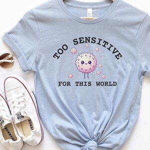 May include: Light blue t-shirt with a graphic of a pink and white cartoon character with the text "Too Sensitive For This World".
