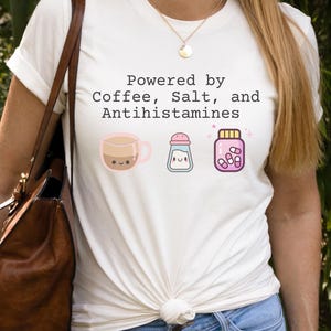May include: A white t-shirt featuring the phrase "Powered by Coffee, Salt, and Antihistamines" with cartoon images of a coffee cup, salt shaker, and pill bottle. The person is wearing a gold necklace and a brown shoulder bag.