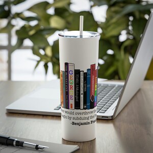 May include: White insulated tumbler with a silver lid and a white straw. The tumbler features a design of colorful book spines with titles like "The Bluest Eye" and "Fahrenheit 451." A quote by Benjamin Franklin is printed below the books.