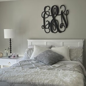 May include: A white bed with a white headboard and a white fluffy comforter. There are several pillows on the bed, including a black and white patterned pillow. A black wooden monogram hangs on the wall above the bed.