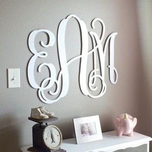 May include: White wooden monogram letters spelling "AM" in a cursive script style, mounted on a light gray wall. The letters are large and decorative, with a delicate, flowing design.