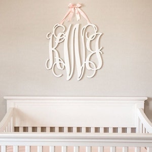 May include: White wooden monogram with the letters K, V, and C in a cursive script, hanging on a light beige wall above a white crib with a white mattress and a pink blanket.