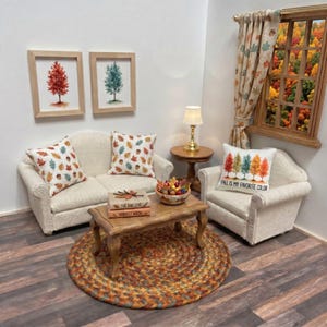 May include: A miniature living room set featuring a cream-colored sofa and armchair with autumn-themed pillows, a wooden coffee table with two books titled The Oak Leaf and Harvest Moon, a bowl of miniature fruit, and a side table with a lamp.