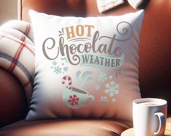 May include: A white square pillow with the words "HOT Chocolate WEATHER" in a decorative font. The pillow features a graphic of a mug with steam, snowflakes, and a candy cane. A white mug of hot chocolate sits on a wooden side table.