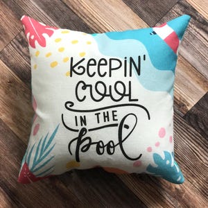 May include: Square decorative pillow with the text "KEEPING COOL IN THE POOL" in black script. The pillow has a white background with colorful tropical leaf and abstract designs in pink, blue, and yellow.