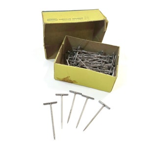 May include: A box of T-shaped pins, also known as T-pins, in a yellow box with the text "OAKVILLE DIVISION" printed on the top. The box is open and the pins are spilling out. There are also several T-pins scattered on a white surface.
