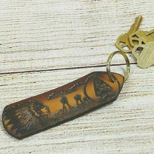 May include: A brown leather keychain with a carved design of a Native American chief and a desert landscape. The keychain has a brass key ring.