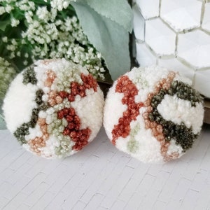 May include: Two round, fluffy pom-poms with a cream-colored base and a pattern of green, brown, and rust-colored sections. The pom-poms are made of a soft, textured material and are likely decorative items or craft supplies.