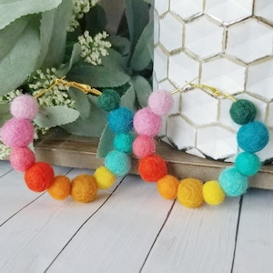 May include: Rainbow-colored felt ball hoop earrings. The earrings feature a gold-colored hoop with a series of felt balls in a gradient of colors, including pink, orange, yellow, green, and blue. The earrings are displayed on a wooden surface.